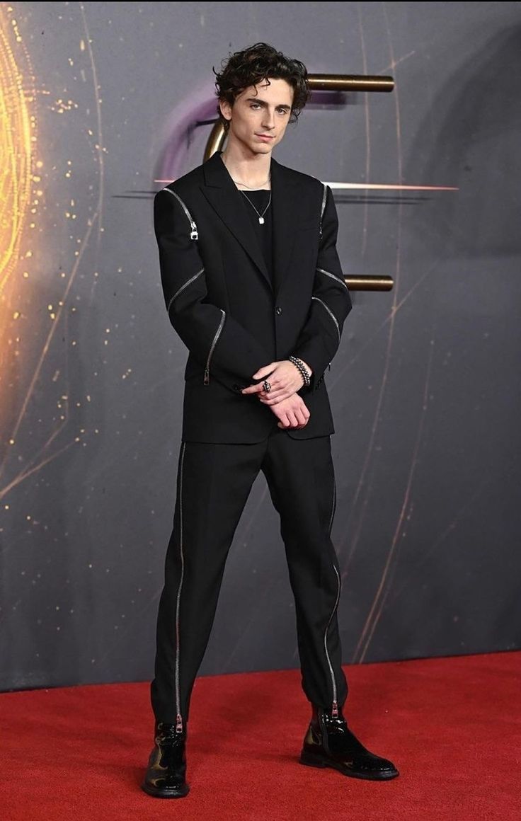 the image shows timothée chalamet standing on a red carpet wearing a black suit. the suit has zipper details on the sleeves and pants. he is wearing black shiny shoes, a necklace, bracelets, and rings. he is looking slightly to the right. the background is a dark color with some golden abstract shapes.