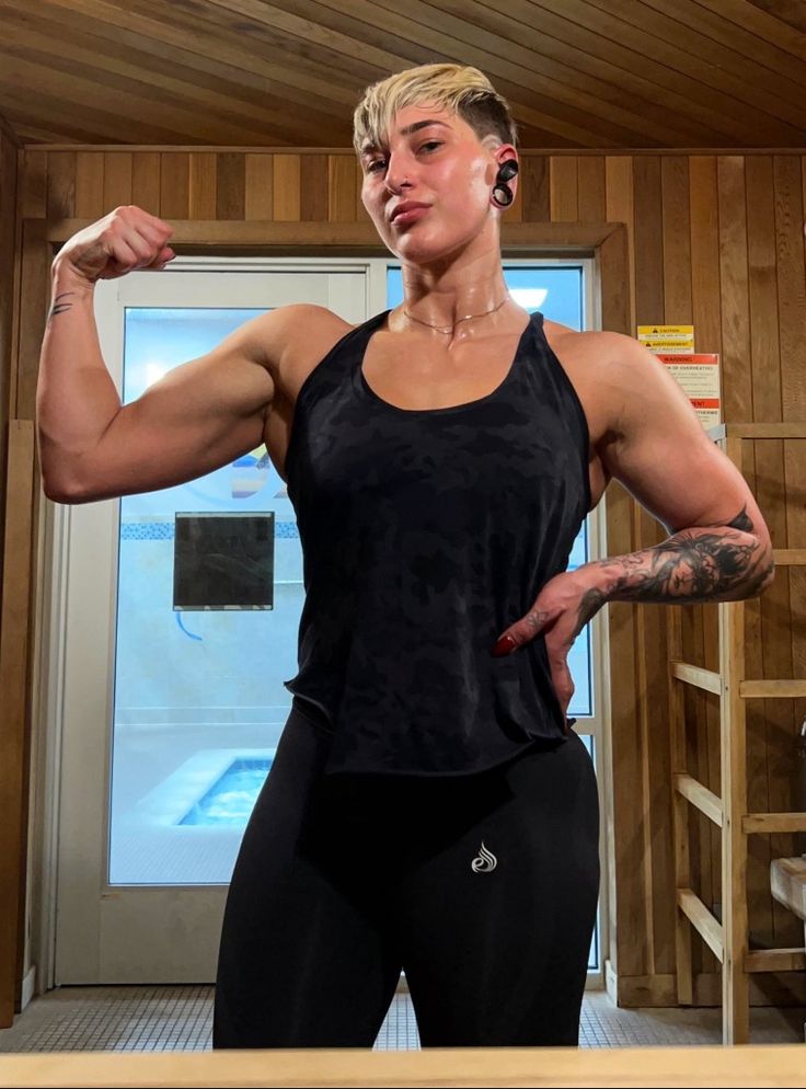 a muscular woman in a black tank top and leggings flexes her bicep. she's standing in a sauna or gym with a woodpaneled interior, a hot tub in the background and a rack of towels and toiletries.