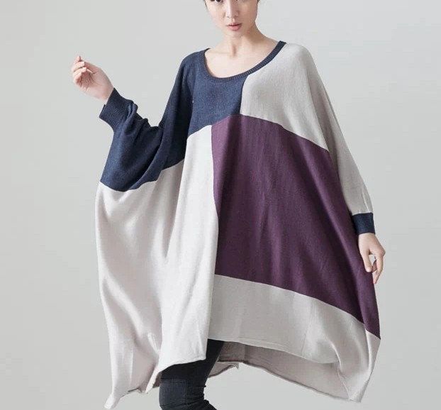 a woman is standing indoors wearing a large, colorblocked sweater. the sweater is a mix of dark blue, purple, and offwhite sections. it has long, wide sleeves. she is wearing dark pants and the background is a neutral grey/white.