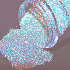a closeup shot of an iridescent glitter jar pouring out. the glitter is a variety of colors but mostly clear and white. the container is a thick glass with a lip and is frosted with the same iridescent glitter. the jar is tilted so that the glitter is pouring out of the opening. the glitter fills the frame and the jar is not fully visible. the glitter is reflecting the light, making it sparkle.