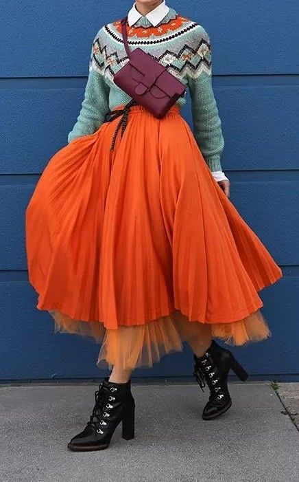 a person is wearing a colorful outfit consisting of a white collared shirt, a light blue and patterned sweater, an orange skirt, and black boots. the person is also wearing a maroon purse that is slung over their chest.