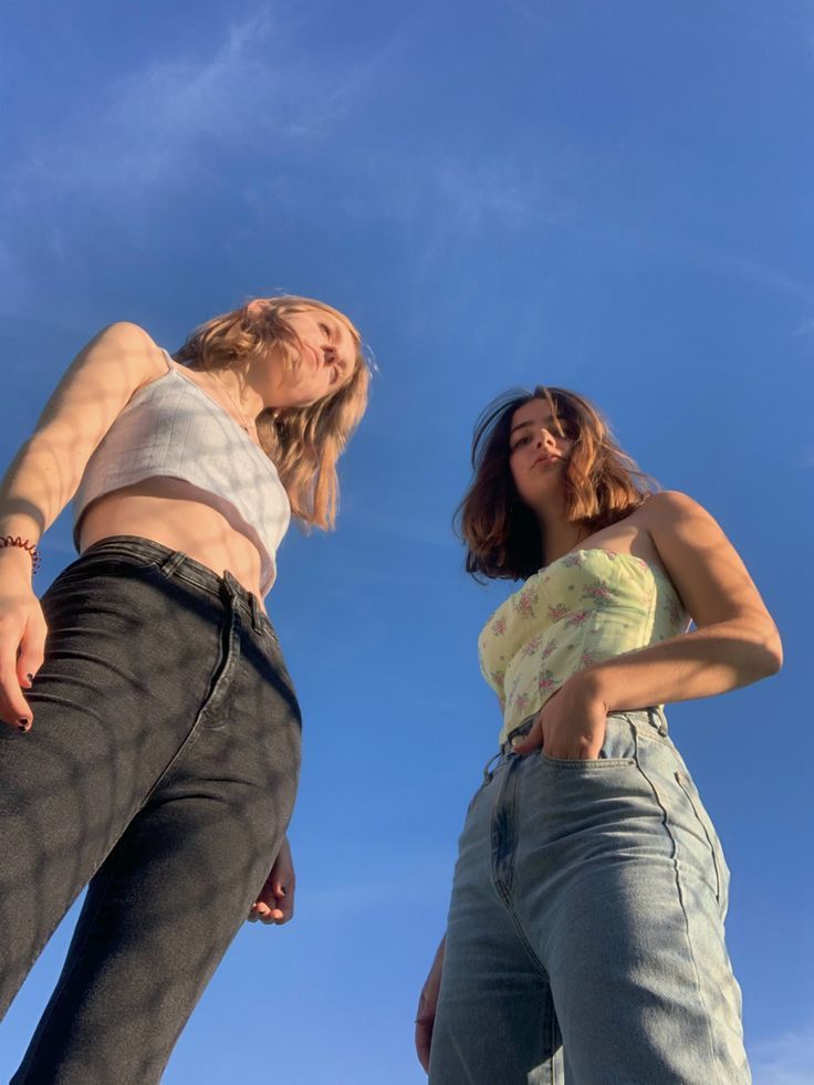 two young women are standing against a bright blue sky, photographed from a low angle. the woman on the left is wearing a white tank top and black jeans, while the woman on the right is wearing a yellow floral top and light wash jeans. they both appear relaxed and casual.