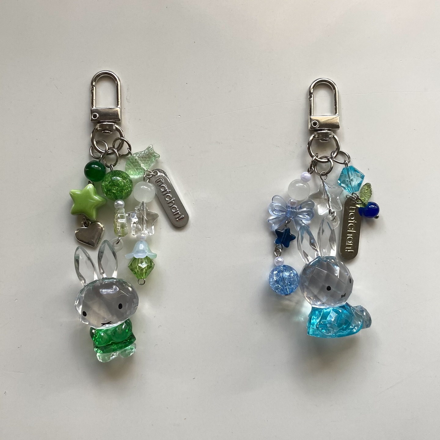 two keychains with crystal rabbit charms are displayed on a white surface. the keychain on the left features green and silver beads and a heart charm, while the keychain on the right features blue beads and a bow. each keychain has a silver clasp and a silver tag with text "hatchan!".