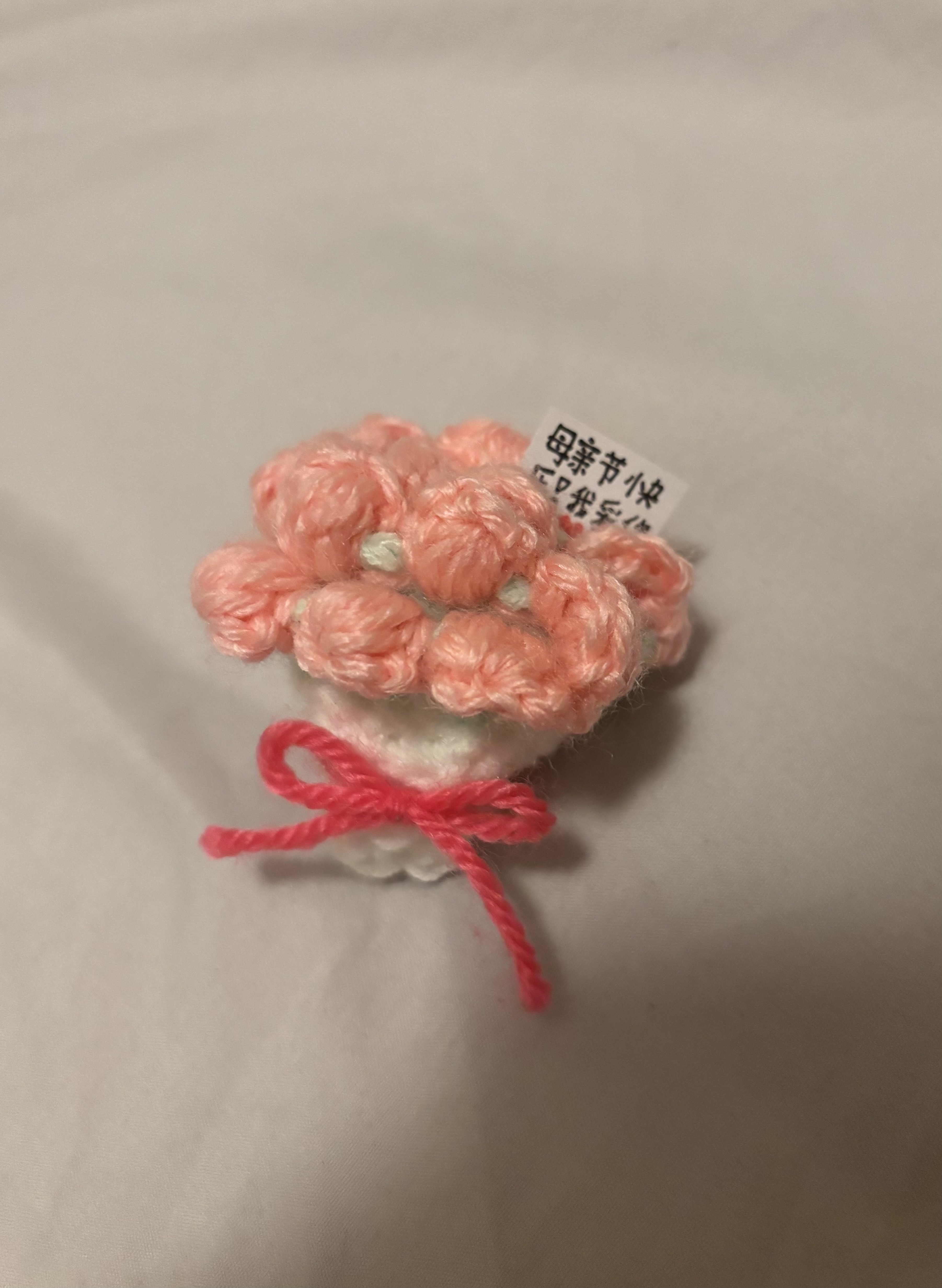 a small, handmade crochet bouquet of pink flowers tied with a pink yarn bow, with a small tag that says "happy mother's day" in chinese. the bouquet is sitting on a white surface.