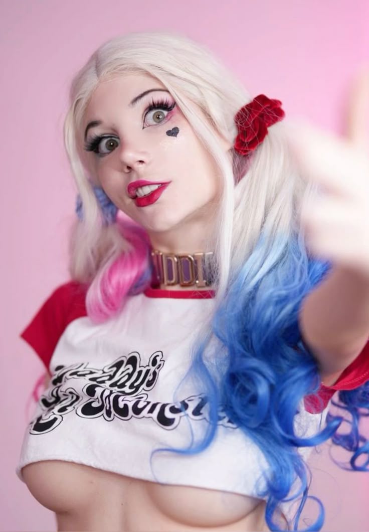 a person dressed as harley quinn from dc comics. she has blonde hair in pigtails with pink and blue dye, a heart tattoo on her cheek, red lipstick, and is wearing a "daddy's lil monster" white crop top with red sleeves and a gold choker. she is posing in front of a pink background while biting her lip and looking at the camera.