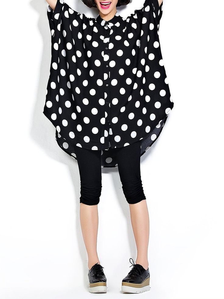 a woman stands in front of a white background wearing a large black and white polka dot shirt, black leggings, and black platform shoes.