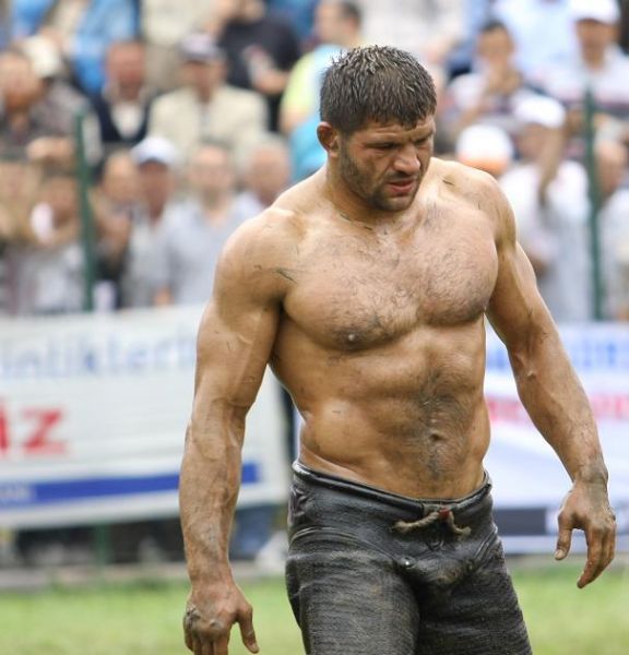 a muscular man with bare chest and mud on his body is standing in a field. he is looking down and has a determined expression on his face. the image is likely of a wrestler and is a portrait of a strong, athletic man.