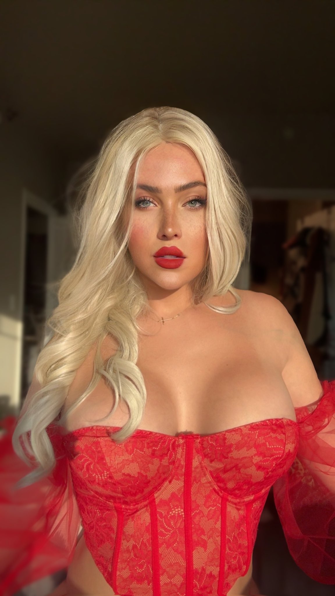 loseup portrait of a blonde woman with full bust wearing a red lace offtheshoulder corset. the woman has long, wavy blonde hair and is wearing red lipstick. the image is welllit and has a glamorous feel.  she is looking directly at the camera. the background is blurred.