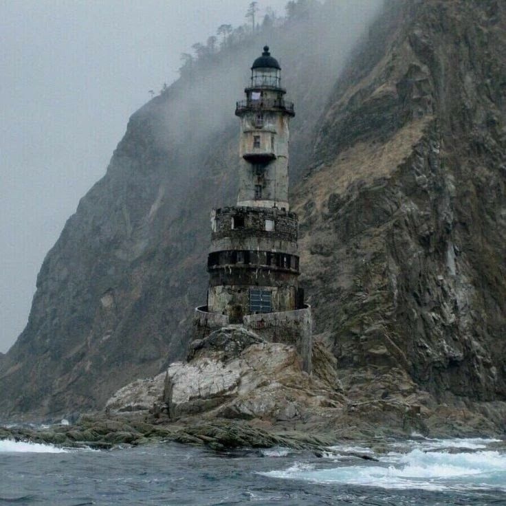 the image shows an old, abandoned lighthouse perched on a rocky island next to a tall cliff. the sea surrounds the base, with waves crashing against the rocks. the sky is overcast and foggy, creating a gloomy atmosphere. the lighthouse is multitiered and made of stone, showing signs of age and disrepair.