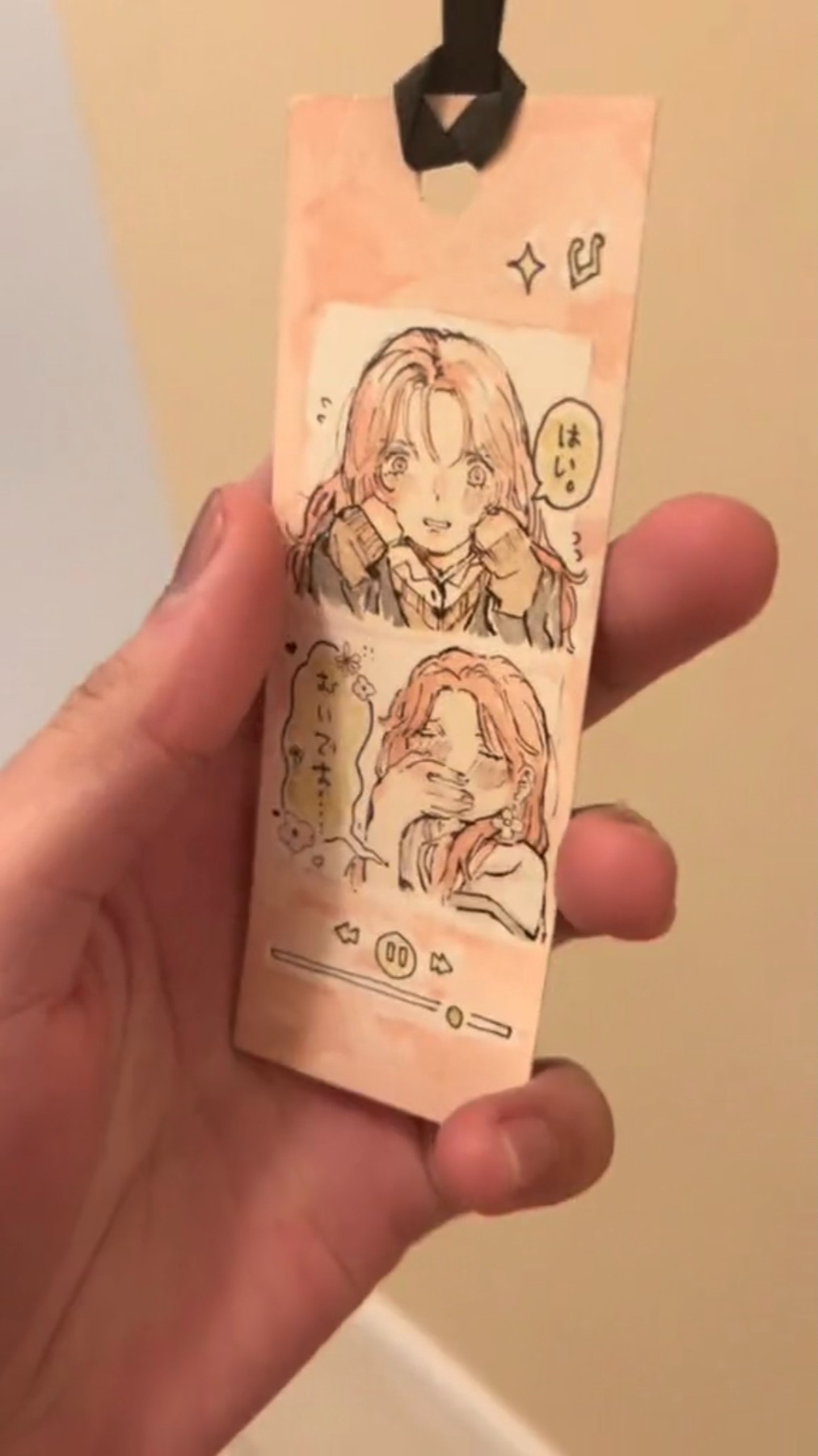 a hand is holding a handmade bookmark. the bookmark is pink and white. the bookmark features an illustration of two girls who are in an anime/manga style. the illustration is on a pink background, and the hand is holding the bookmark against a light brown background. the girls are wearing pink and white clothing, and they have long hair and bright eyes. the bookmark appears to be a hand drawn illustration.