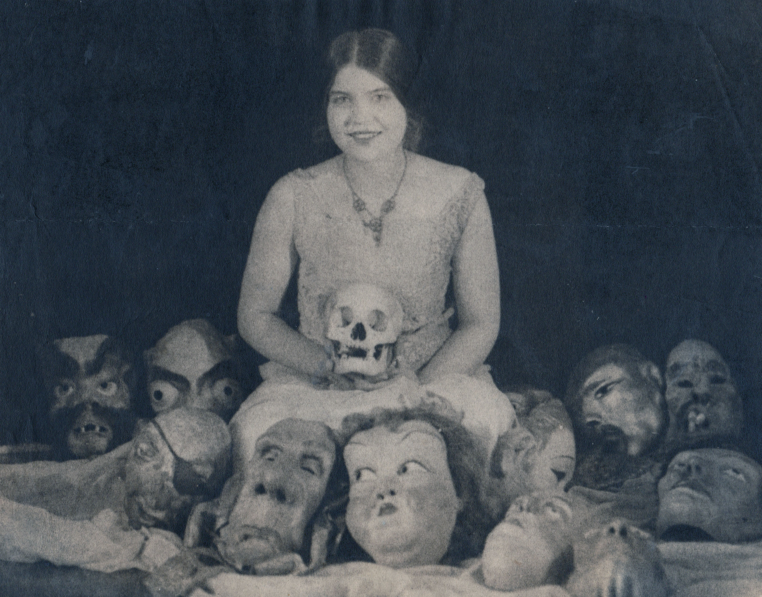 a vintage black and white photograph shows a young woman sitting amidst a collection of masks and a skull. she is dressed in a lightcolored dress and has dark hair parted in the middle. her expression is calm and she holds a skull in her lap. the masks surrounding her are various grotesque and monstrous faces. the overall tone of the image is dark and mysterious, with a hint of gothic horror. the photo's style and subject matter suggest it might be from the 1920s or 1930s.