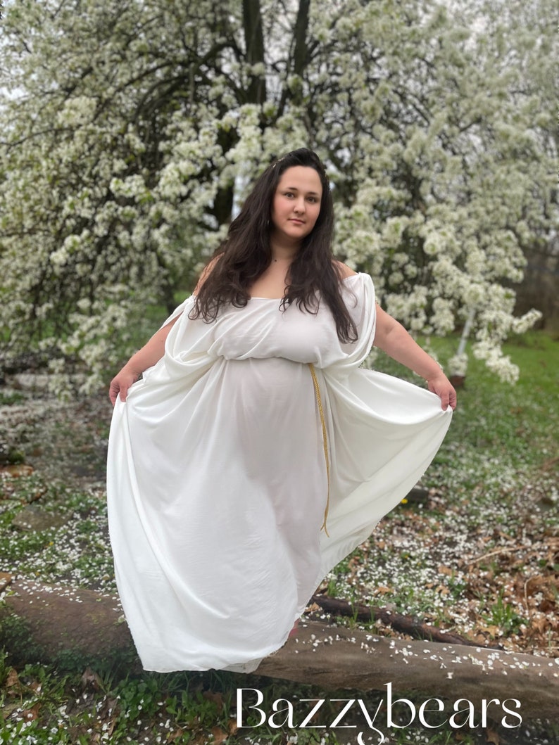 a woman in a flowing white grecianstyle dress stands in a park surrounded by blooming cherry trees. the dress is offtheshoulder with a long skirt that pools around her feet.  she is barefoot. the ground is covered in fallen petals. the watermark "bazz ybears" is visible in the bottom of the image.