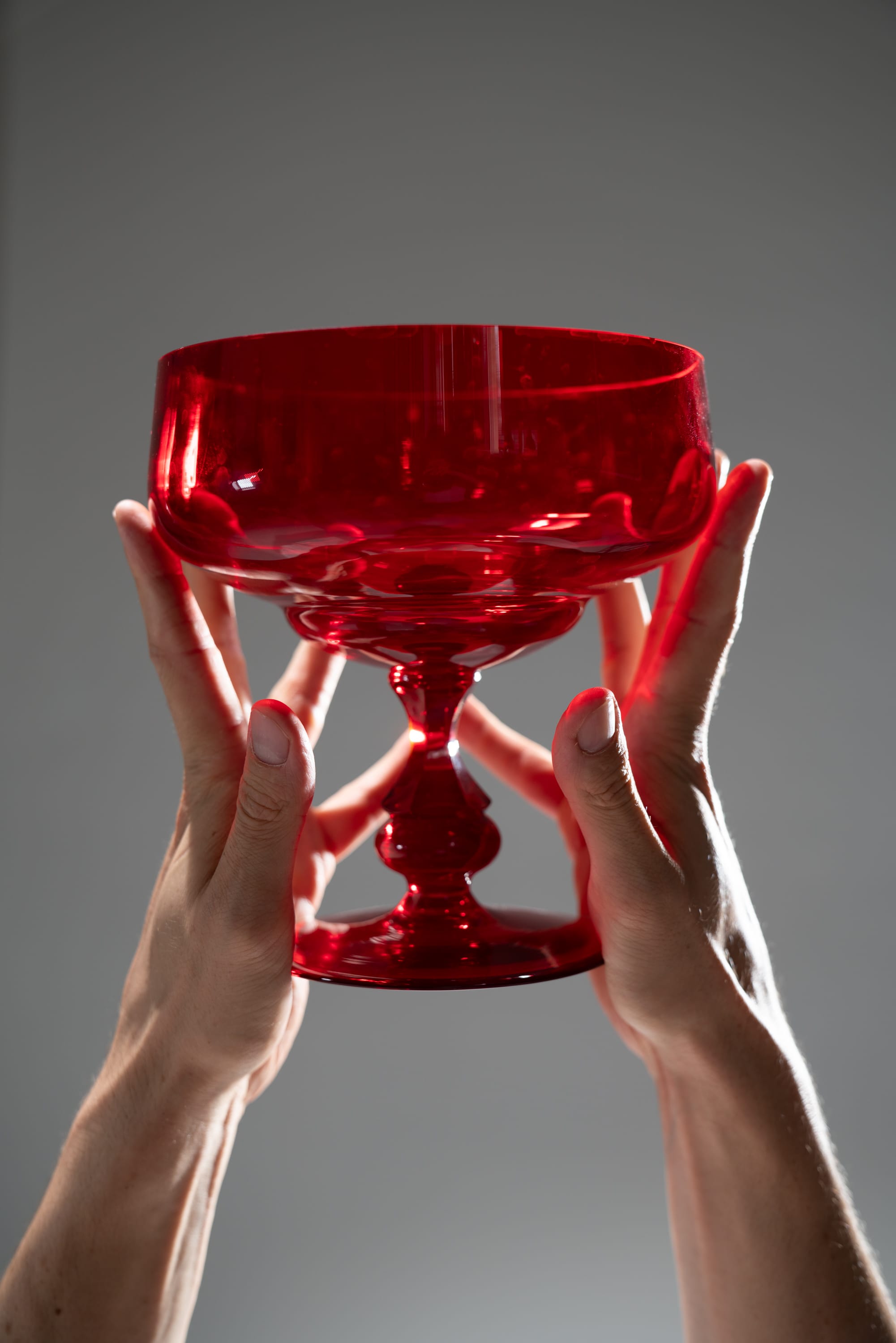 two hands are holding a red glass. the glass has a wide bowl and a slender stem with a round base. the light is shining through the glass, making it appear translucent.