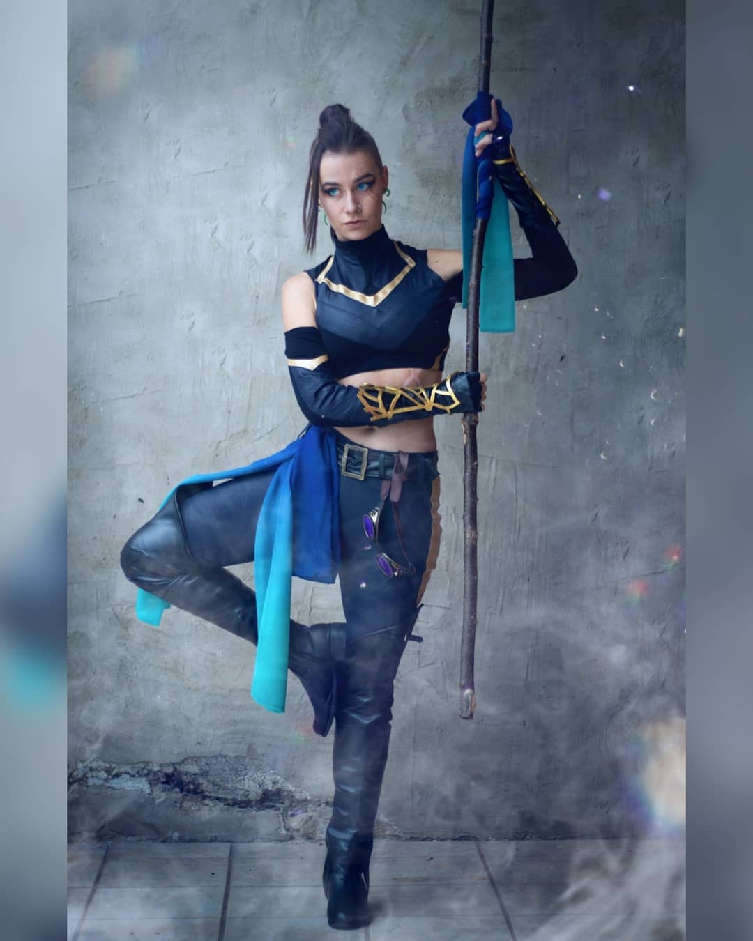 the image showcases a female cosplayer portraying a fantasy warrior. she is dressed in a meticulously crafted costume featuring a darkcolored top, leather pants, and high boots. a flowing teal and blue fabric drapes from her waist, adding a touch of elegance to her otherwise fierce attire. the cosplayer holds a staff, suggesting a character wielding magical abilities. her pose is dynamic, with one leg raised, and her expression is intense and focused. the background is a textured grey wall, and subtle smoke or mist adds a sense of depth and drama to the image. the overall aesthetic blends fantasy elements with a modern feel, highlighted by the cosplayer's stylish attire and the professional quality of the photograph.