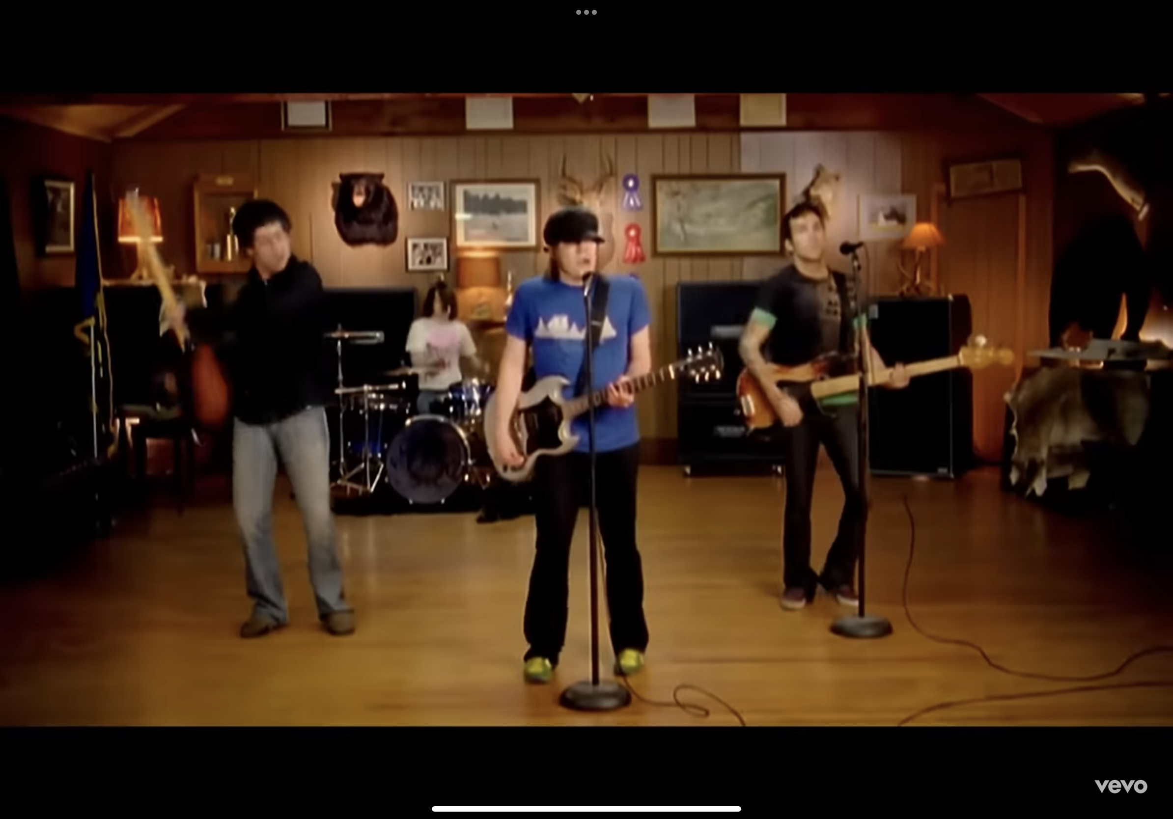 a still image from what appears to be a 2000s era music video. four young men are playing instruments on a wooden floor in a lodgelike setting. the walls are woodpaneled and decorated with taxidermied animal heads and framed awards. the lighting is bright and artificial. the overall aesthetic suggests a rock or pop punk band.