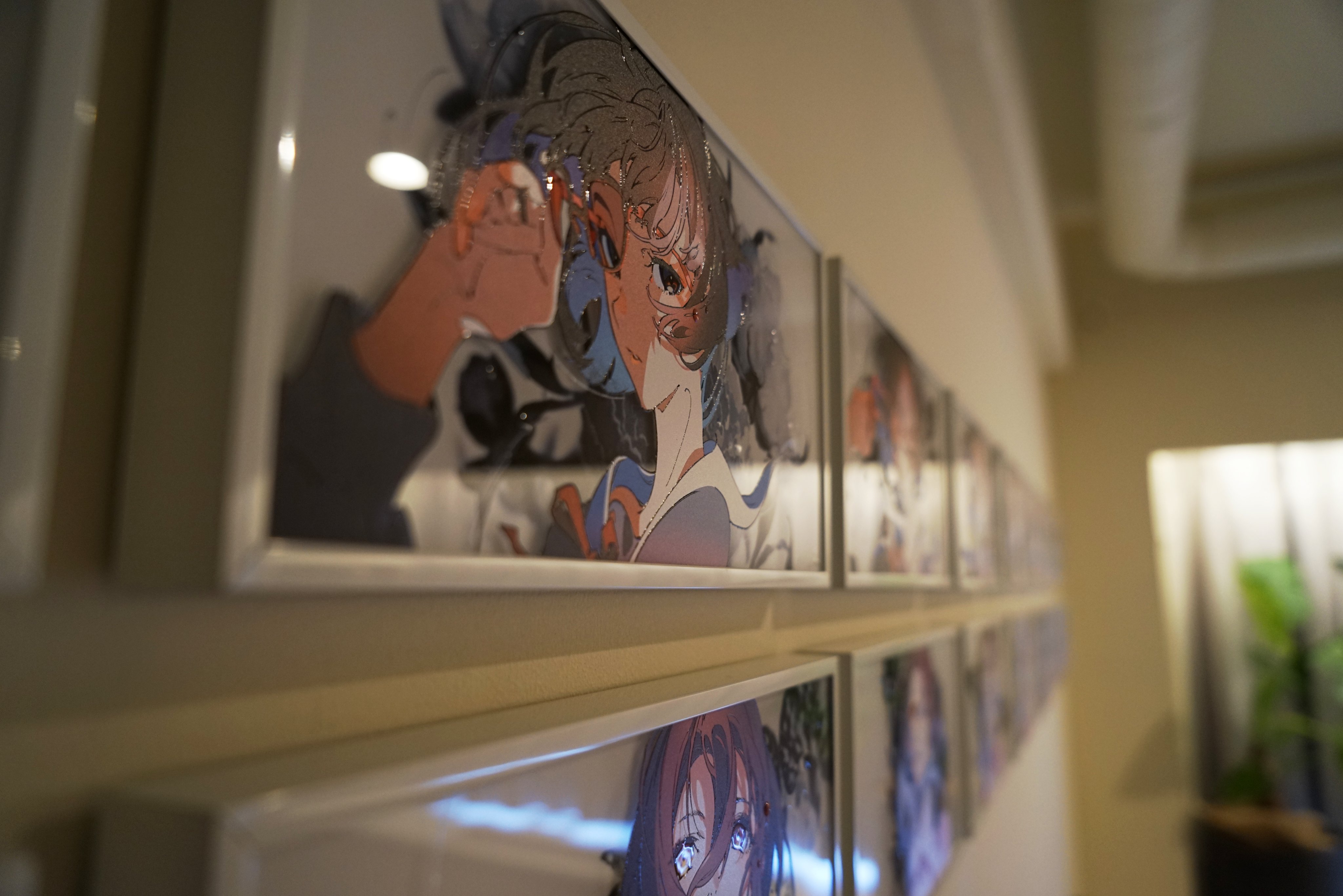 a row of animestyle drawings are framed and hung on a wall.  the drawings feature female characters, and the frames are white.  the wall is creamcolored, and there's some greenery visible in the background on the right side. the lighting is bright and natural.
