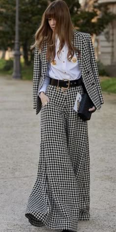 a young woman is wearing a stylish black and white gingham blazer with wide leg pants. she is wearing a white shirt underneath the blazer and a black leather belt around her waist. the woman's hair is styled in long, soft waves. she is carrying a black clutch bag. she is looking down, giving the impression of a casual and stylish look.