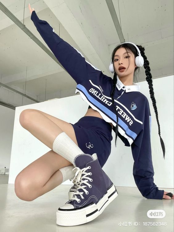 a young asian woman poses for a fashion shot. she is wearing a sportychic outfit, including a blue and white cropped sweatshirt, a matching skirt, white socks, and blue and white converse sneakers. she has long black braids and is wearing white headphones. the background is a white wall with a blue stripe. the image has a modern and trendy feel.