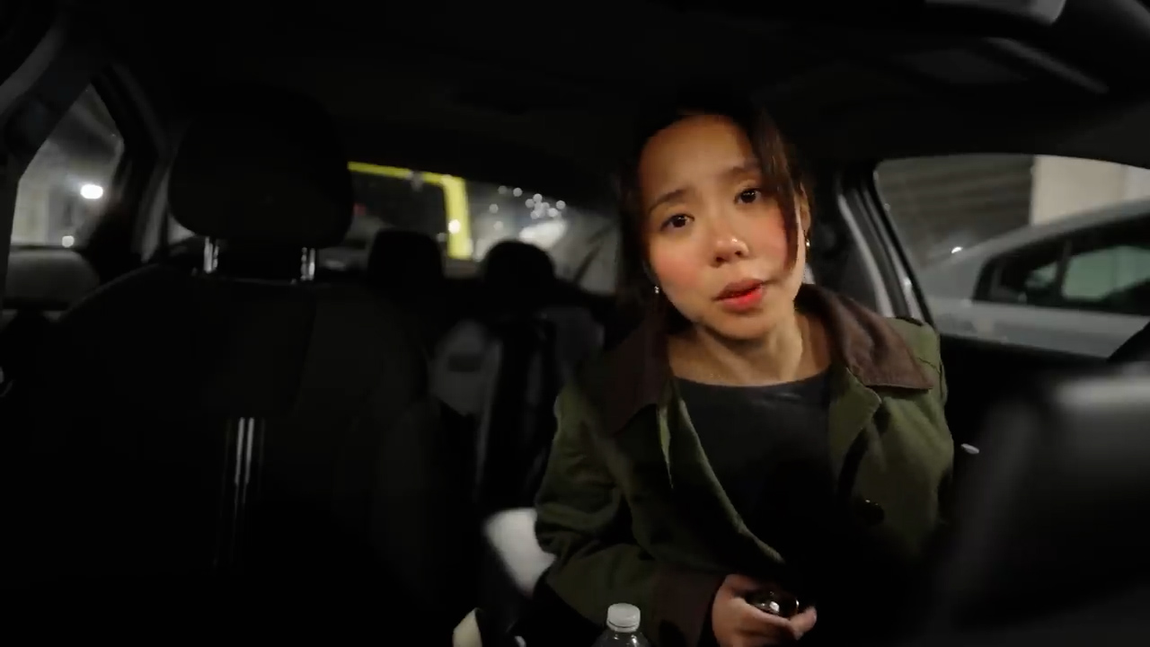a young woman with dark hair and a green jacket is sitting in the driver's seat of a car. she is looking at the camera with a thoughtful expression on her face. the car is parked in a parking garage, and the background is dark.