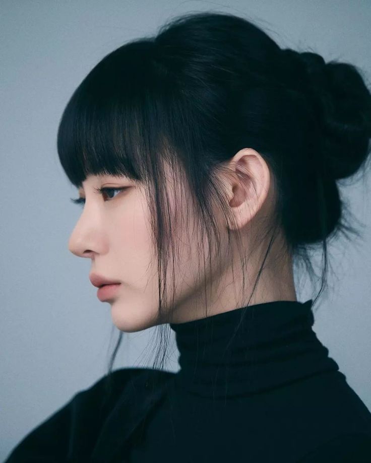 a closeup profile shot of an asian woman with black hair pulled back into a bun. she has blunt bangs and loose strands framing her face. she is wearing a black turtleneck sweater and has a serious expression. the background is a neutral color.