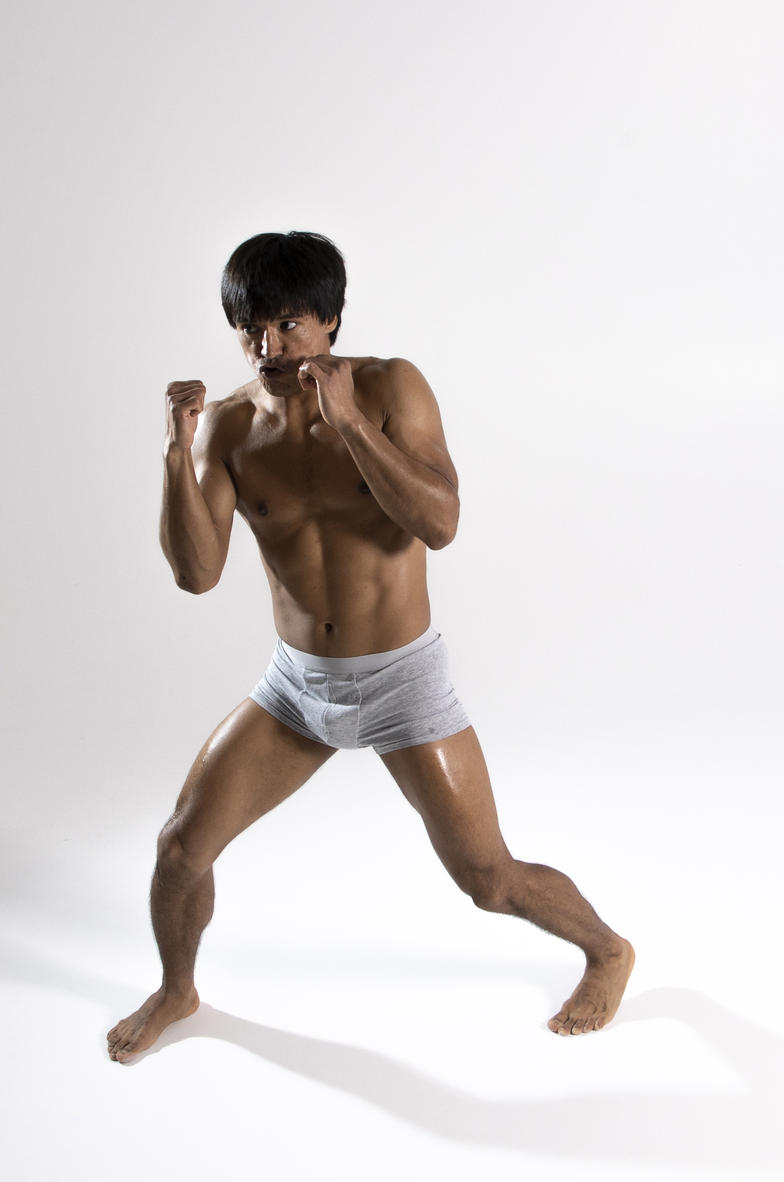 a shirtless man is in a boxing stance wearing grey underwear. his skin looks oiled. he's barefoot and looking directly at the camera. his shadow is behind him.