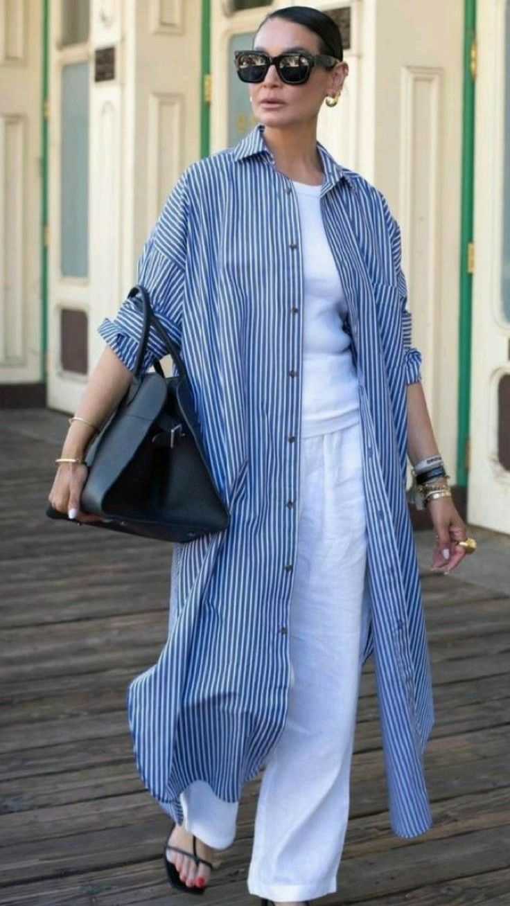 a stylish woman in a blue and white striped shirt, white pants, and black sunglasses poses on a city sidewalk. she is carrying a black handbag and accessorizing with jewelry.