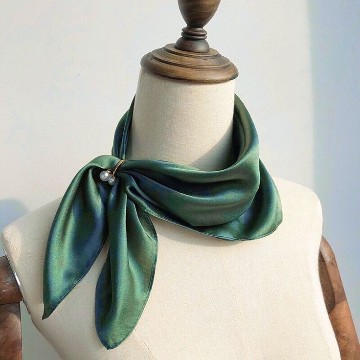 loseup shot of a dark green silk scarf draped around a mannequin's neck. the scarf is a lustrous satin material and is tied in a simple knot. a small, delicate pearl brooch is pinned to the scarf, adding a touch of elegance. the mannequin is light beige and has a wooden base. the background is a plain, light grey, making the scarf the focal point of the image.  the overall style is sophisticated and minimalist.