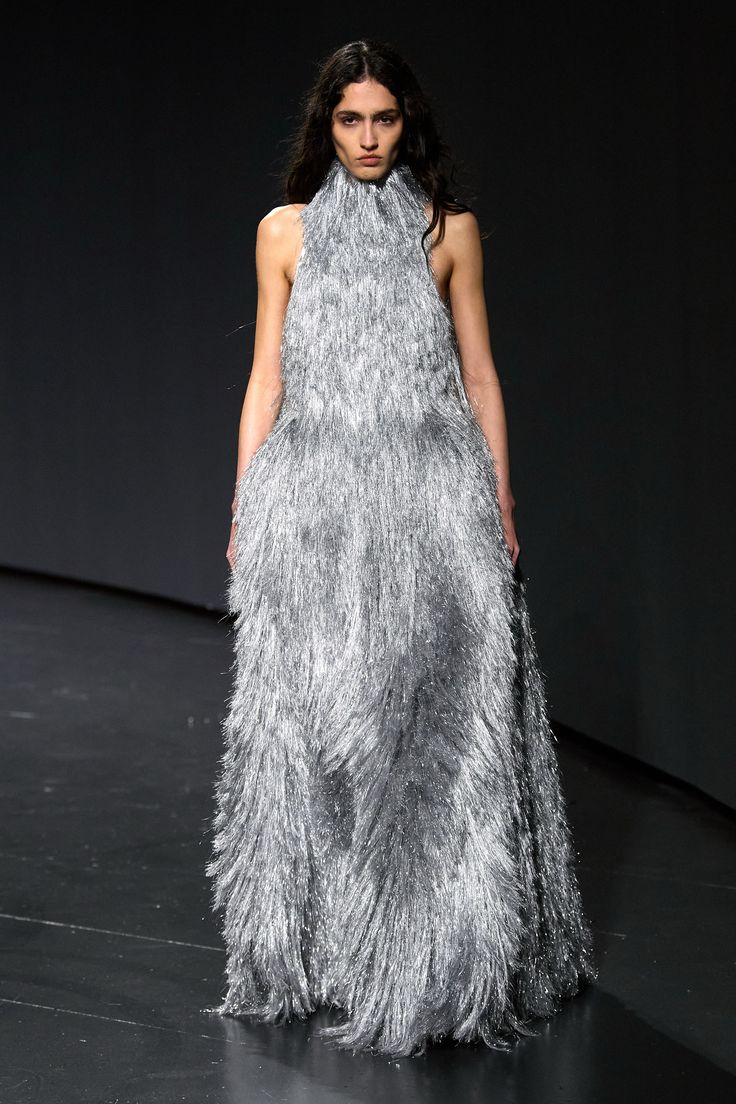 a model is walking on a runway wearing a long, silver gown. the dress has a high neck and is covered in tinsel. the model has long, dark hair and is looking straight ahead. the background is dark.