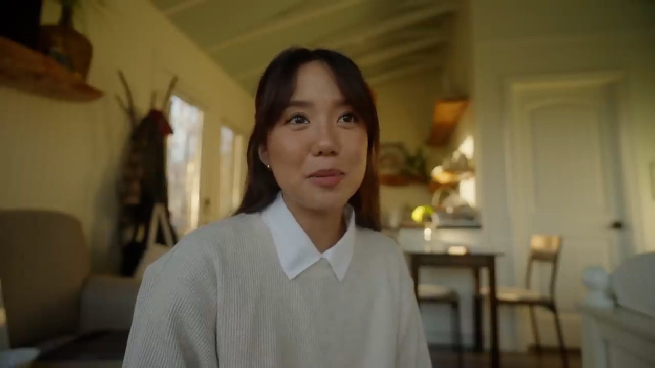 a young asian woman sits in a cozy living room, smiling warmly. the room is filled with natural light streaming through a window, highlighting the warm and inviting atmosphere.