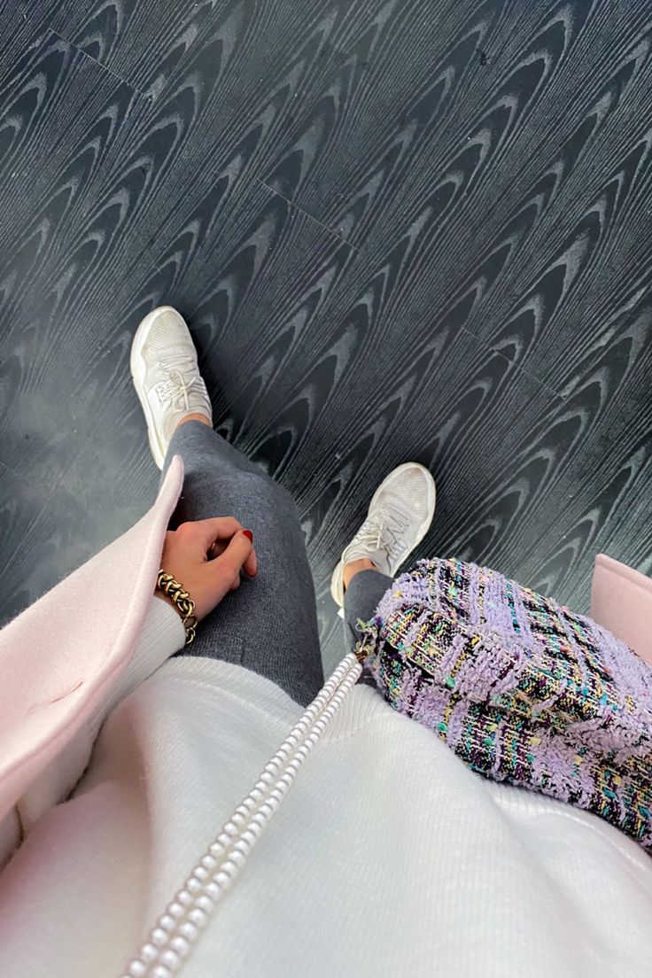 a highangle, closeup shot of a woman's outfit. she's wearing white sneakers, grey pants, a light pink coat, a white sweater and a colorful tweed bag with a pearl necklace. the floor is dark and textured. the image is focused on the clothing and accessories, showcasing a stylish and trendy outfit.