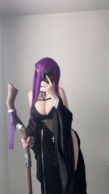 a person with purple hair, presumably a wig, is dressed in a cosplay outfit consisting of a black dress with a low neckline, a robe, and a cross necklace. they are taking a selfie with their phone, obscuring their face. in their left hand, they hold a staff with decorative markings. the setting appears to be indoors against a plain white wall.