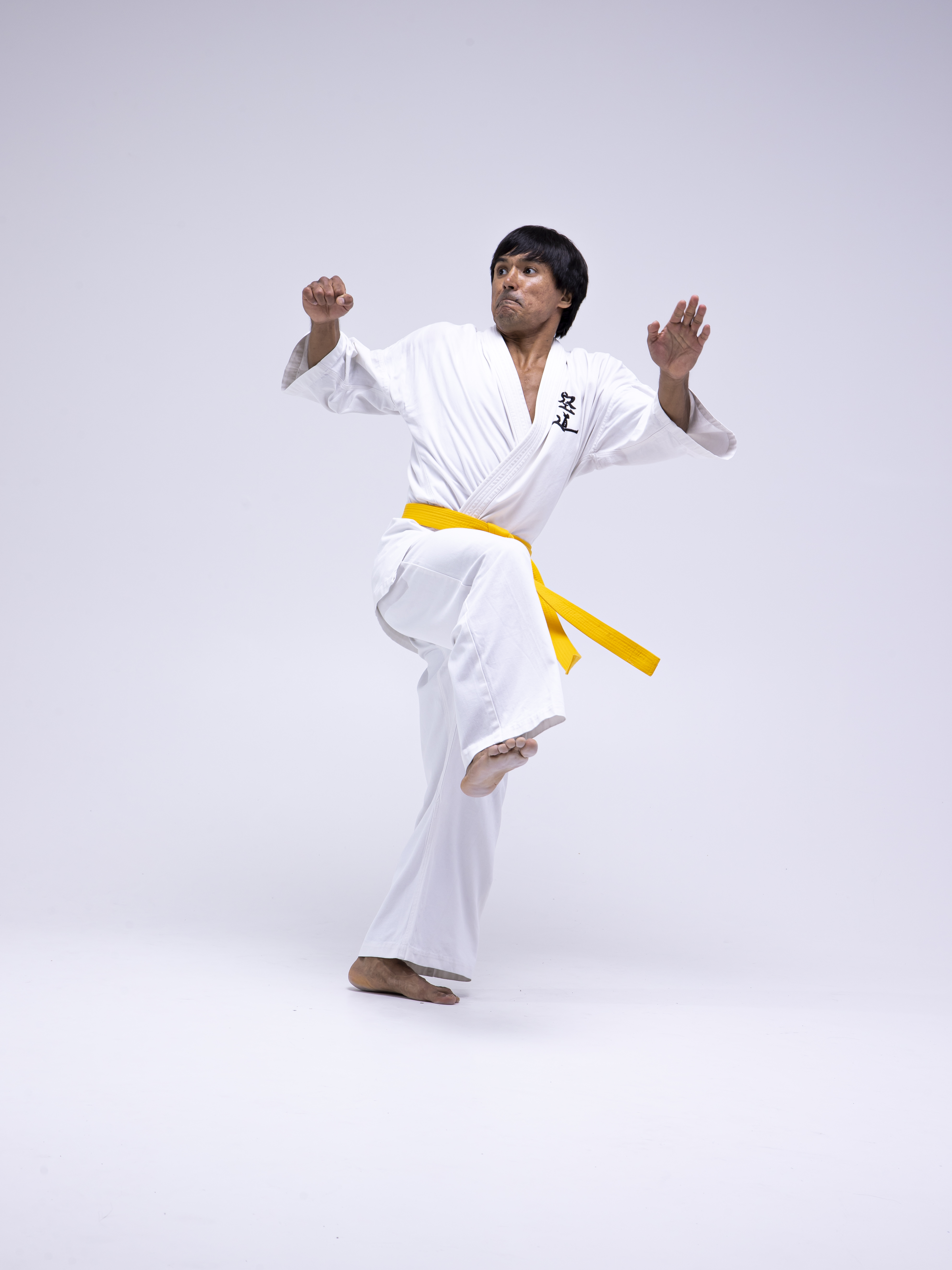 a man in a white karate uniform with a yellow belt is striking a martial arts pose in a studio shot indoors. he is barefoot and has one leg raised.