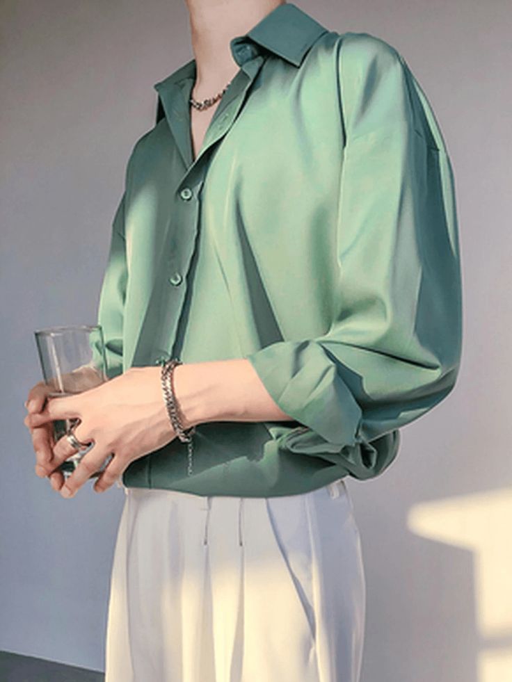 a man wearing a green longsleeved shirt, white pants, a silver chain necklace, a silver bracelet, and a silver ring on his finger. he is holding a glass of water in one hand and is posing for the camera.