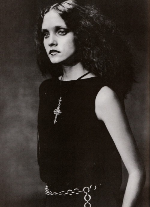 a black and white portrait of a woman with long dark hair, wearing a black dress, a cross necklace, and a chain belt. she has a serious expression and stares directly at the camera. the image is in a vintage style.