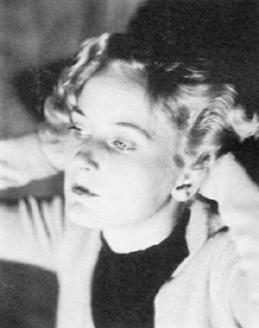 the image is a black and white closeup portrait of a young woman. she appears to be from the 1920s or 1930s, judging by her hairstyle and clothing. her hair is styled in loose, wavy curls, and she's wearing a lightcolored, possibly knitted, cardigan or sweater.  her expression is soft and somewhat pensive; her gaze is slightly downcast. the focus is soft, adding to the overall vintage and artistic feel of the photograph. the lighting is gentle and creates a subtle shadowing around her features. the background is blurred and indistinct, drawing attention to the subject.