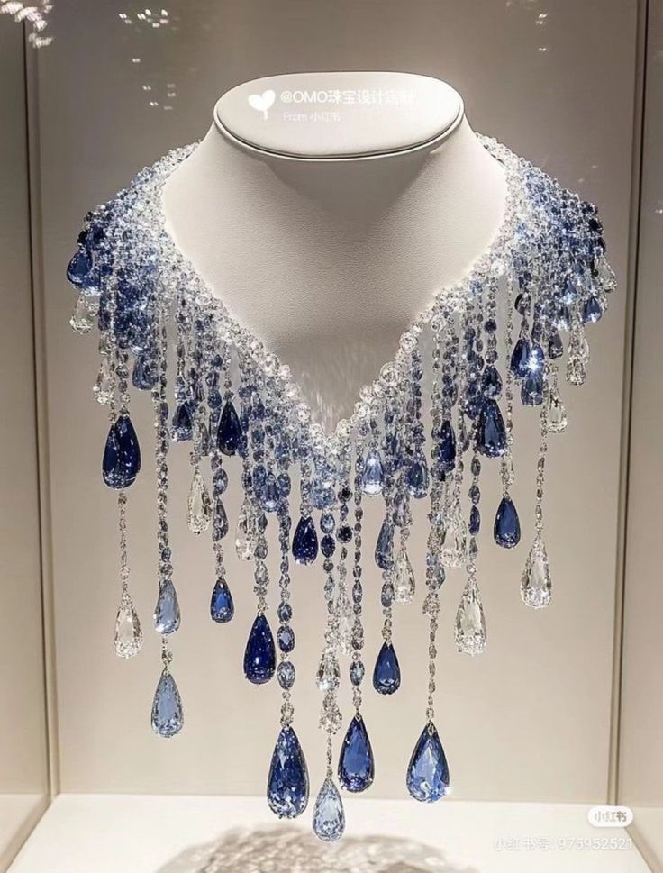 a stunning necklace is displayed on a white mannequin. the necklace features a gradient of blue and clear gemstones, with numerous strands of crystals cascading down. the overall design exudes luxury and elegance.