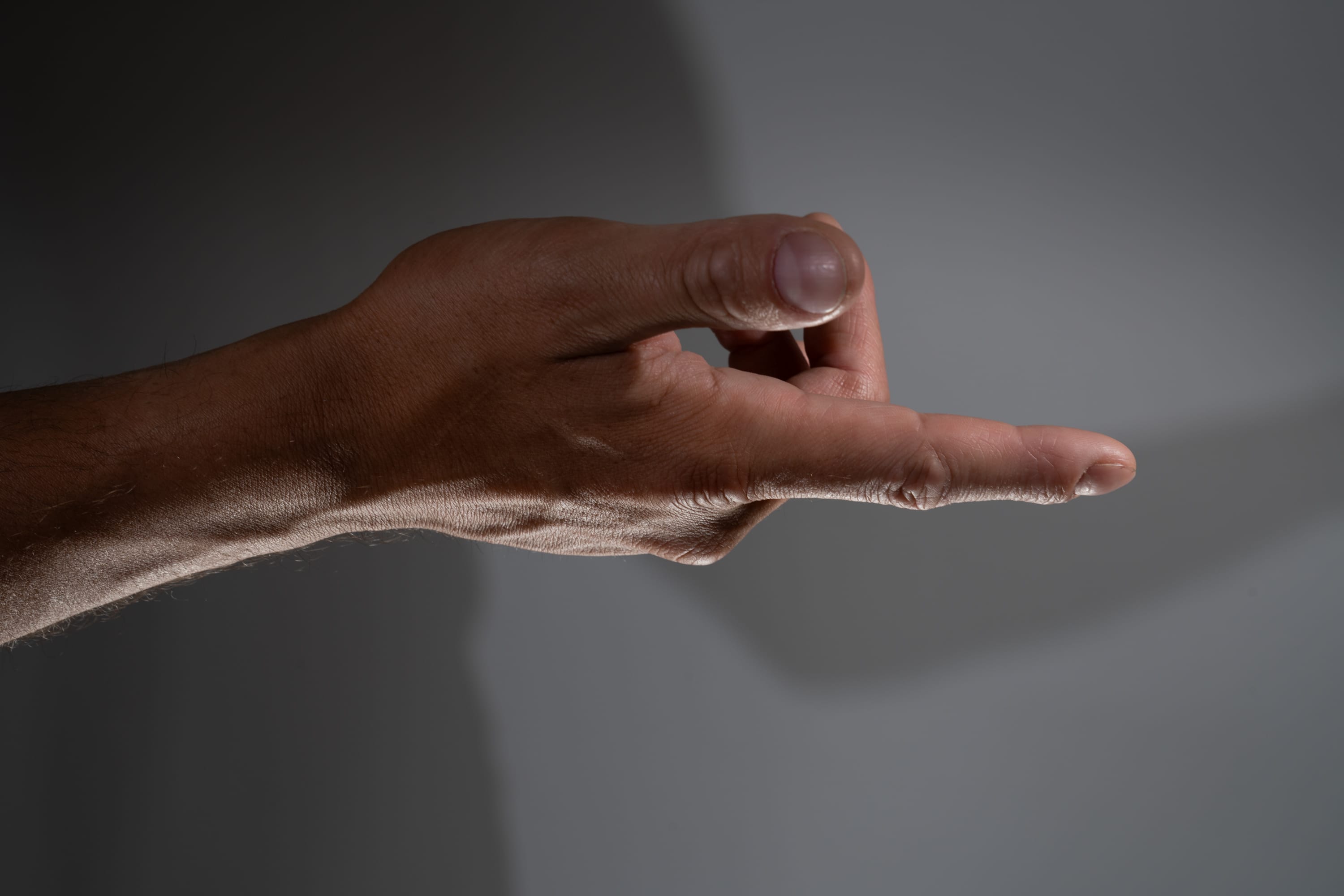 the image shows a male hand with mediumdark skin tone extended towards the right side of the frame. the hand is making a gesture where the index finger is extended, and the remaining fingers are curled into the palm. the background is a neutral gray tone, and the image is lit with side lighting, creating a distinct shadow of the hand on the background.
