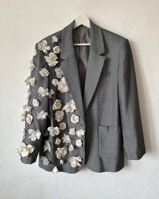 a grey blazer with white floral embellishments on the left side, hanging on a white hanger against a white wall.