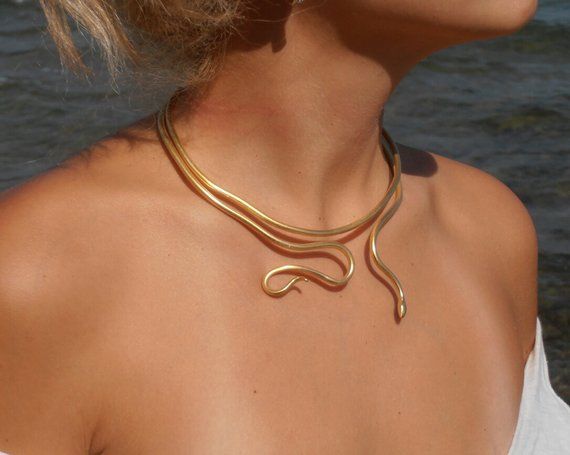 a woman is wearing a gold necklace that resembles a snake. she is standing in front of a body of water.