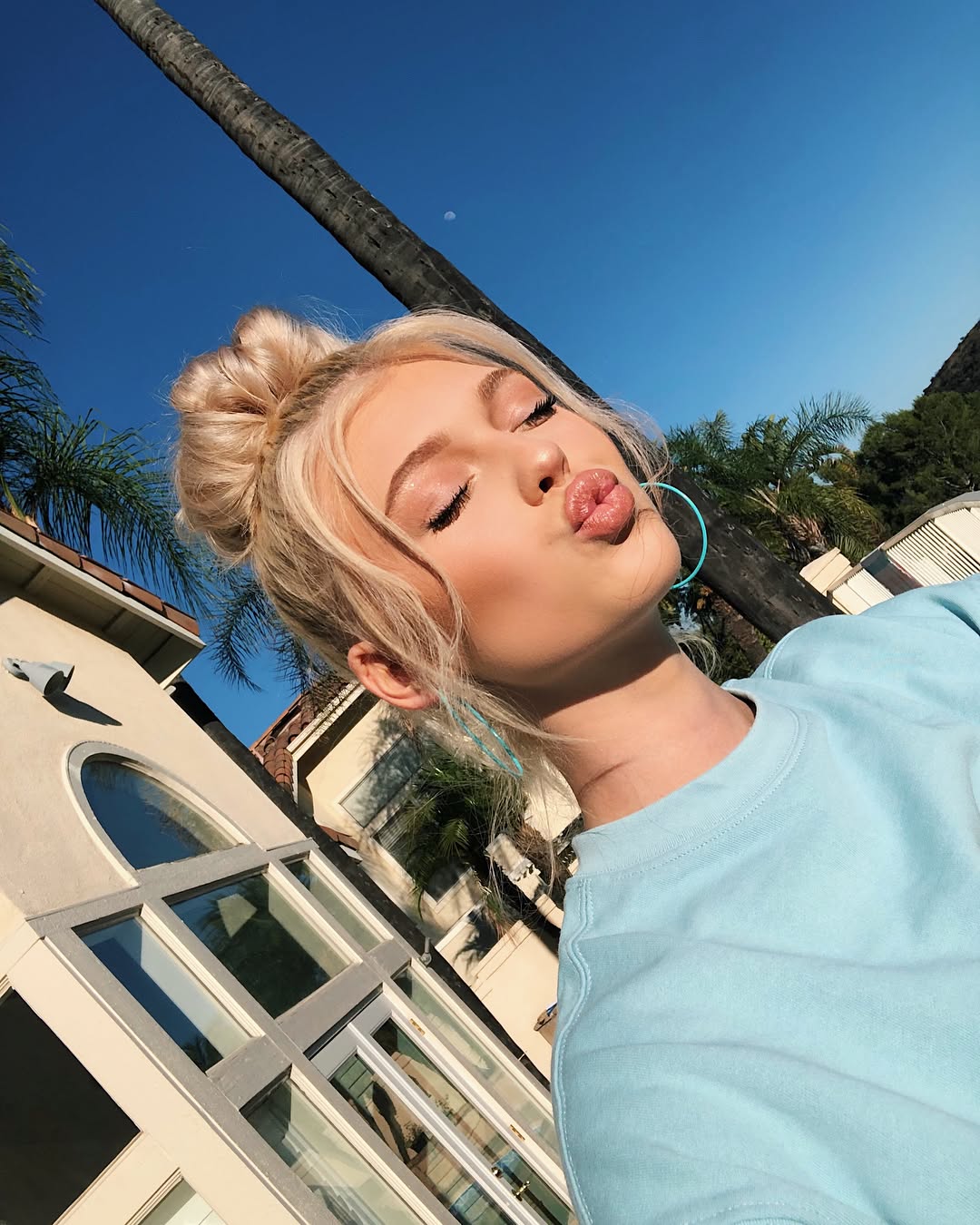 a woman with blonde hair in a bun is pouting with her eyes closed. she is wearing a blue sweatshirt and hoop earrings. the photo is taken outdoors during the daytime, with trees and buildings in the background.