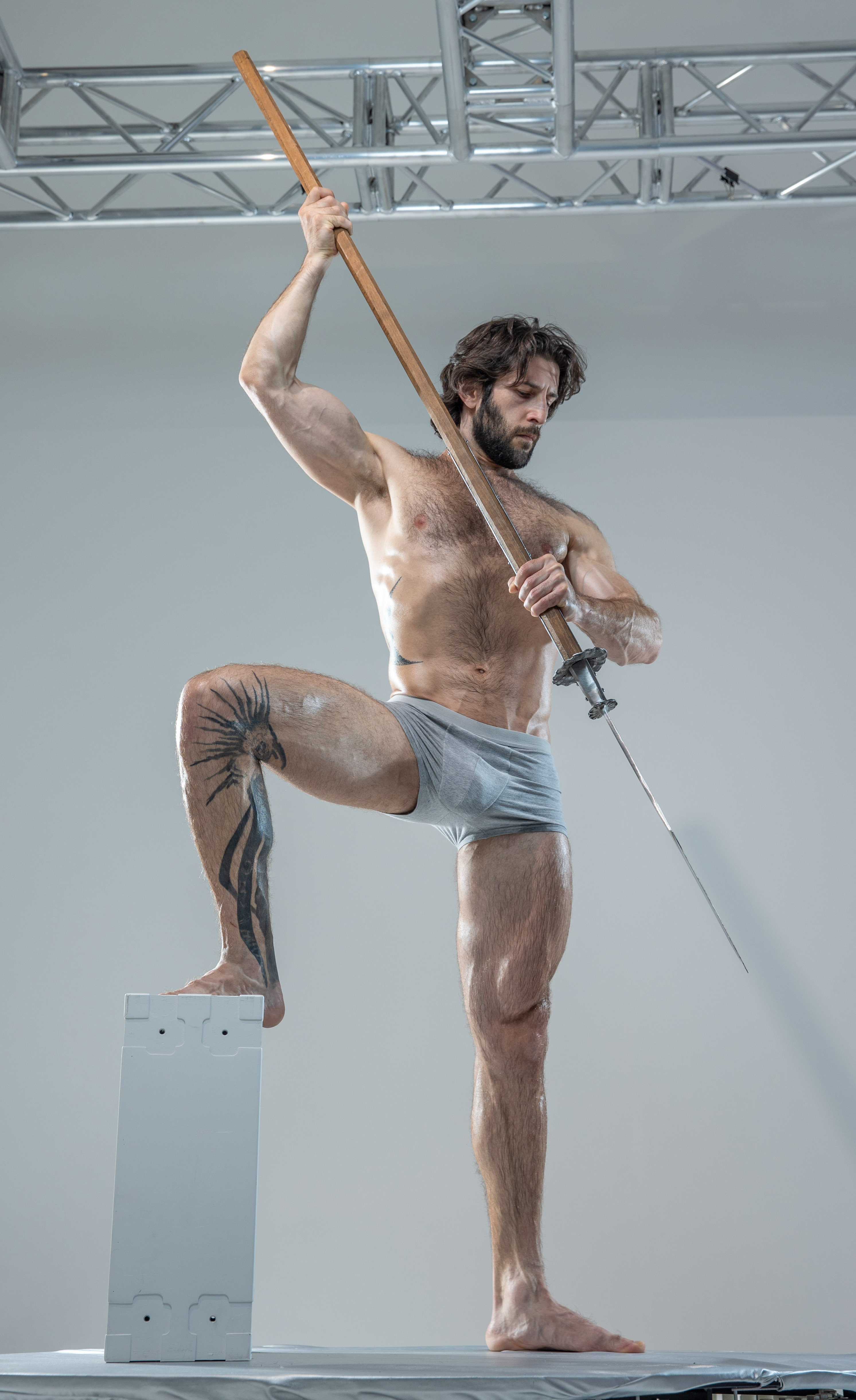 a muscular man with long hair and a beard stands on a white platform in a studio setting. he is holding a spear aloft in a dynamic pose, showcasing his physique. the image is taken from a low angle, emphasizing his strength and power. he is wearing only grey underwear and has a tattoo on his left leg.  the studio has a metal structure visible in the background.
