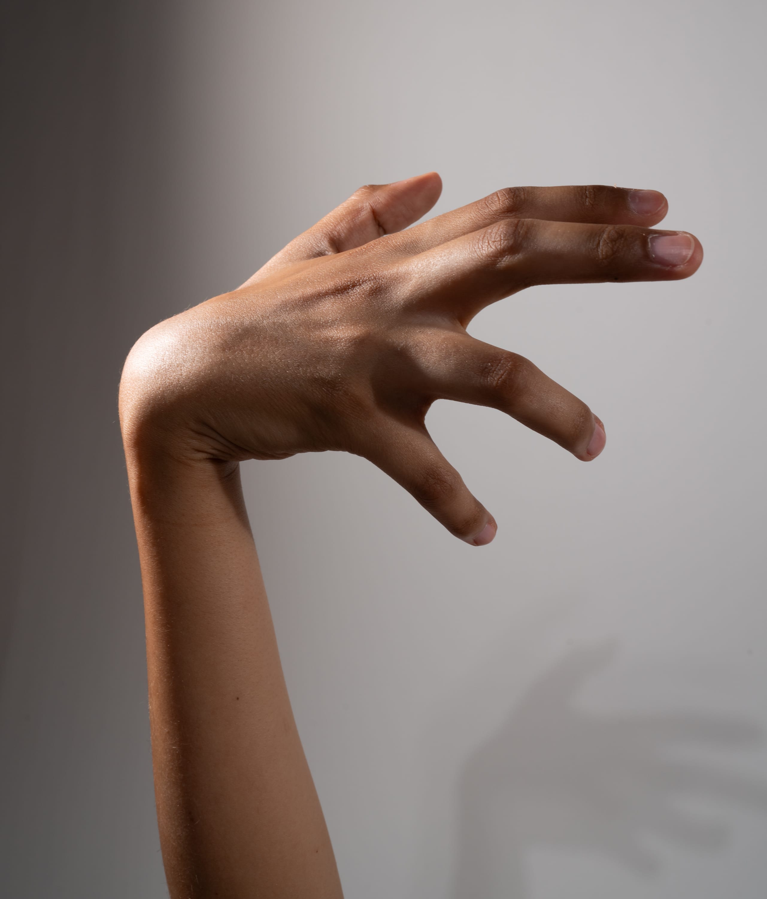 a left human arm and hand extended, with fingers curled in a clawlike gesture, against a gray background. the hand is illuminated by studio lighting, creating a dramatic shadow to the right of the arm.