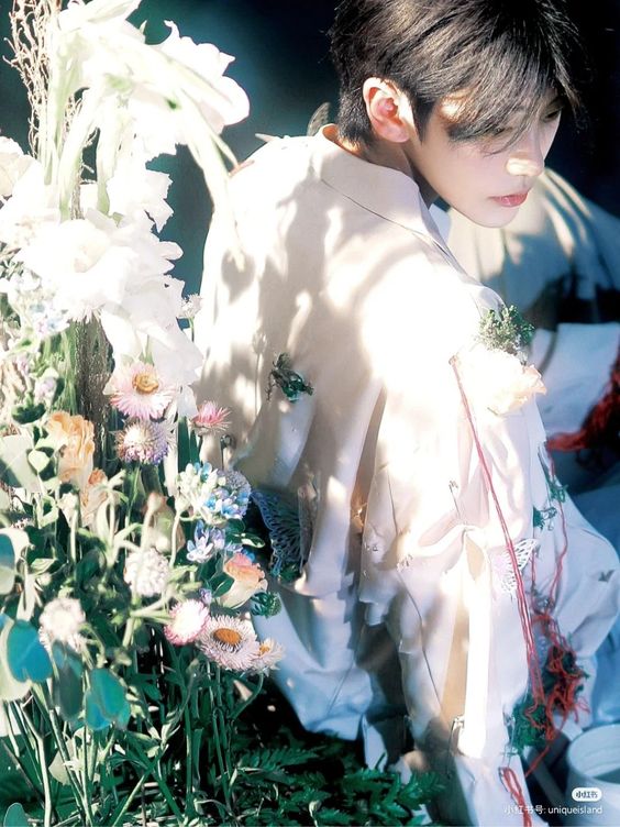 he image shows an asian man in a white, flowy, embellished top. he's positioned in front of a large display of flowers in various shades of white, blue, and yellow. he's looking down, and his hair is styled with a fringe that covers his forehead. the lighting is soft and diffused, creating a gentle and dreamy atmosphere.