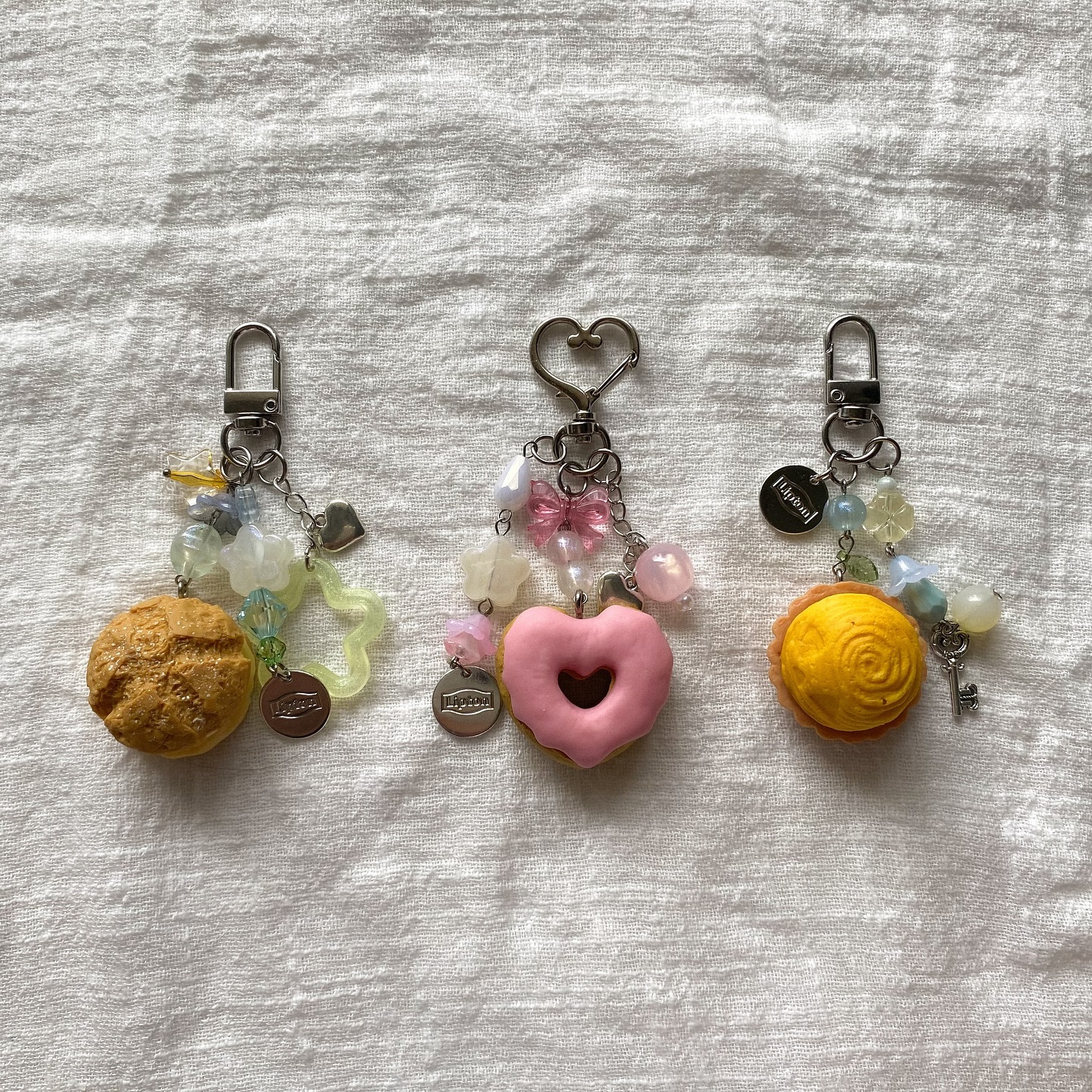 three handmade keychains lie on a white textured surface. the keychains are topped with metal clasps of different shapes (dshape and heartshape), connected to miniature charms resembling food items and adorned with pastelcolored beads and decorative elements such as bows and stars. each keychain features a different food charm: a textured round bread, a pink heartshaped donut, and a spiral patterned pastry. additional charms include metal tags labeled "lipton" and a small key. the color palette consists primarily of pastel hues, adding to the overall aesthetic of the accessories.