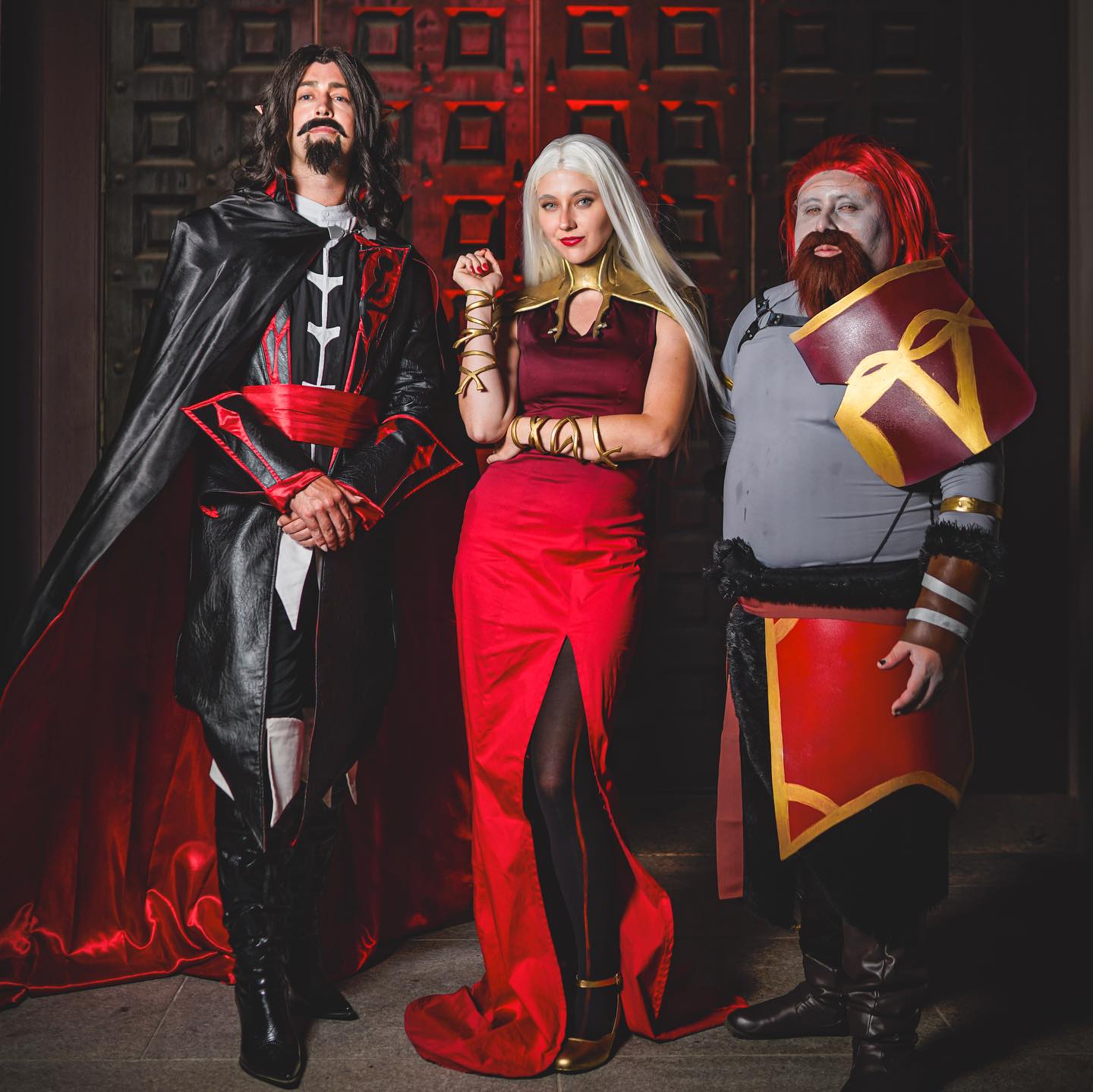 a captivating group cosplay portrait featuring three individuals in elaborate costumes. the central figure is a woman dressed as a queen, adorned in a flowing red gown with a thighhigh slit, gold accessories, and long blonde hair. to her left stands a man portraying a vampire, in a black leatherlike coat with red accents, and a serious expression. to her right, a man in a grey tunic and red warriorlike armor. the backdrop is a dark doorway with red accents adding to the dramatic atmosphere.  the costumes are highly detailed and the overall image suggests a dark fantasy setting.