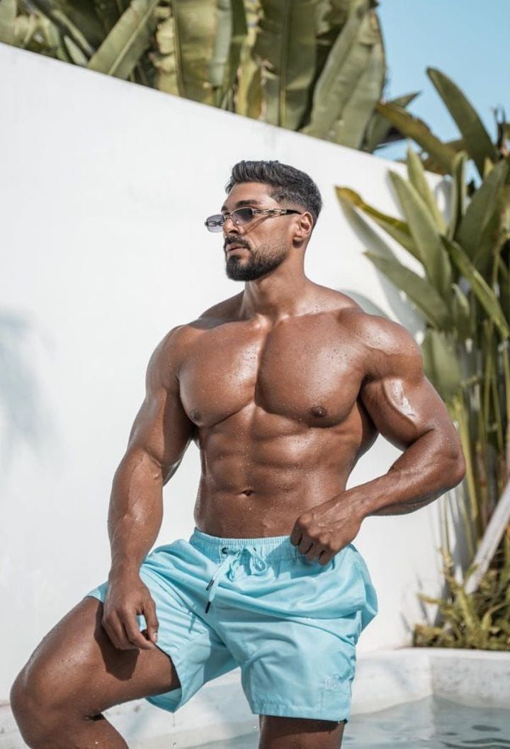 the image showcases a muscular, tanned man with a welldefined physique. he's wearing light blue swim trunks and sunglasses. the setting appears to be outdoors, poolside, with tropical plants and a white wall in the background. the man is posing, showcasing his muscular build. the overall mood is one of summer, relaxation, and health. the image likely aims to promote swimwear or a healthy lifestyle.