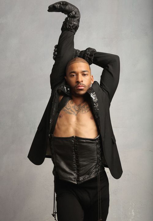 a man wearing all black, a jacket, leather gloves, a corset, and pants is posing in a studio. he is in a dramatic pose with his arms extended above his head.