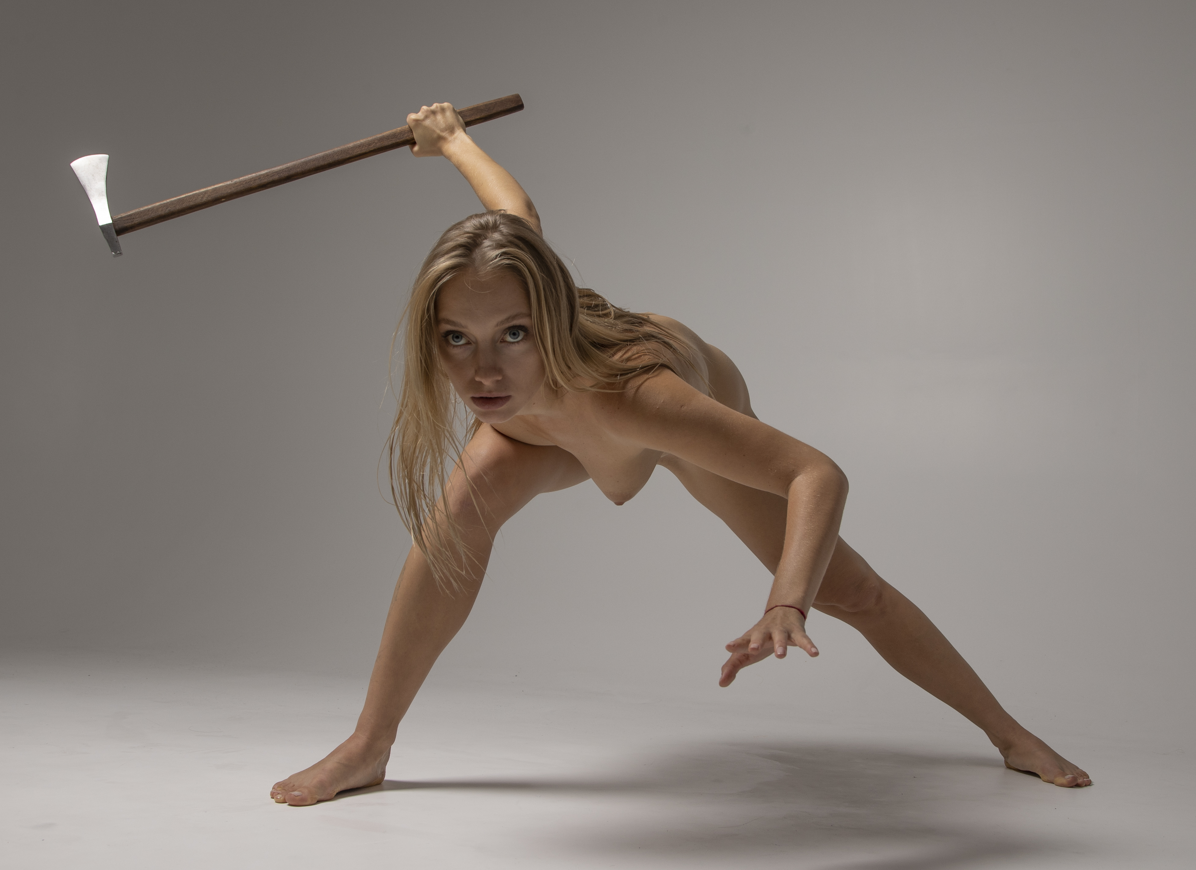 a nude woman is standing with legs spread and bent at the knees. she is holding an axe over her head with both hands. she is looking at the camera.