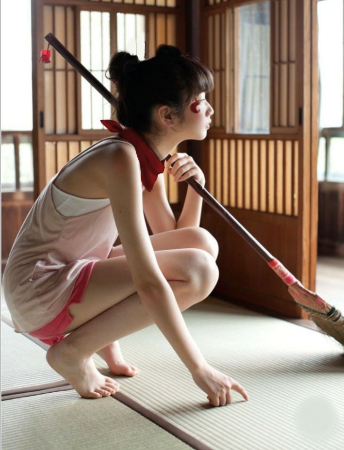 a young asian woman in japaneseinspired clothing is crouching on tatami mats indoors. she is barefoot and wearing a red bandana around her neck. she has red markings on her face. she is holding a broom. the background includes a wooden screen or door.
