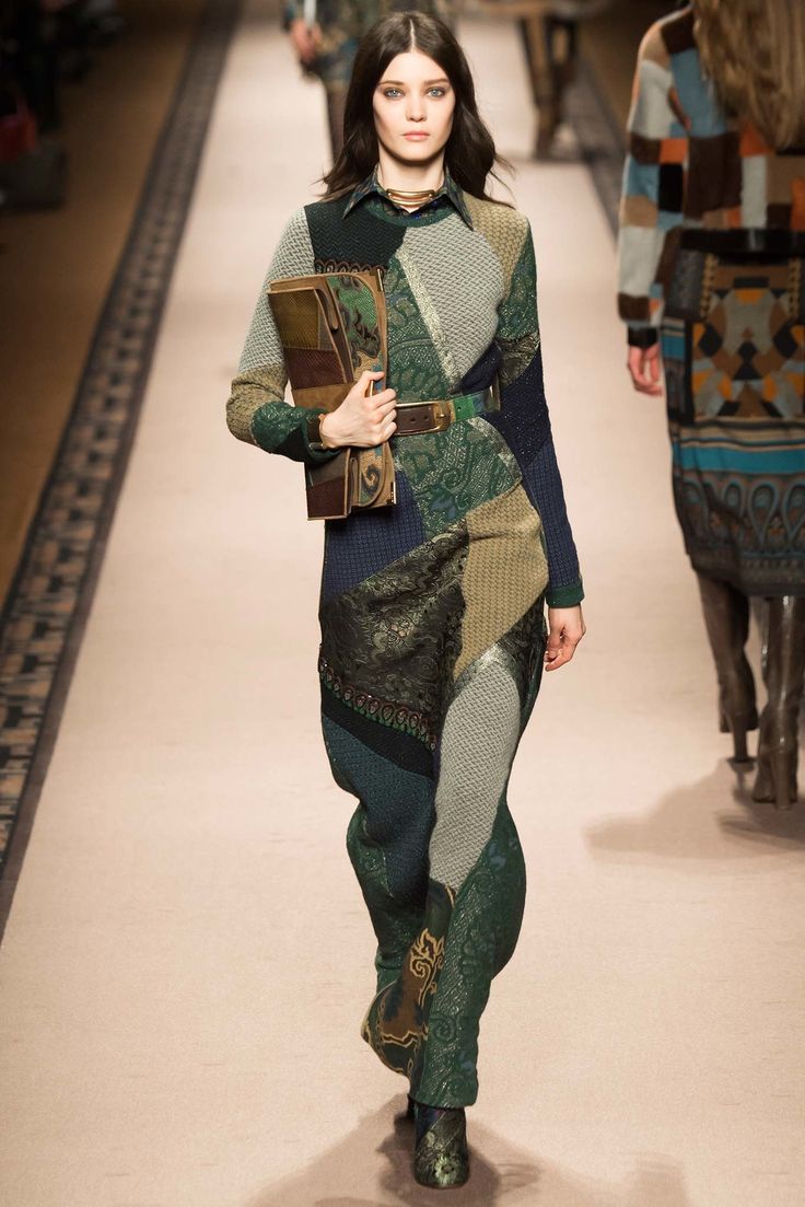 a model walks the runway wearing a long, patchwork dress with paisley and other patterns in shades of green, brown, and beige. the dress is made of different fabrics pieced together. she carries a matching patchwork handbag and wears highheeled ankle boots. the background shows a runway with other models and a light beige floor. the overall style is bohemian and fall/winter appropriate.